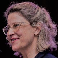 A woman with light blonde, wavy hair and glasses smiles while speaking at a microphone. She has a warm expression and is dressed in a dark jacket. The background features soft, colorful lighting