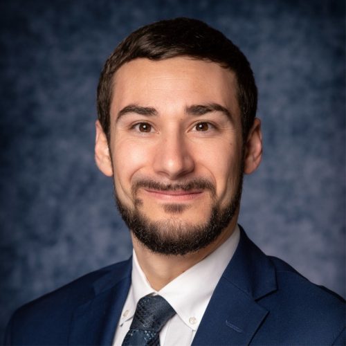 A man with a beard and short hair smiles at the camera, wearing a dark suit and a light-colored shirt with a tie. The background is a textured blue