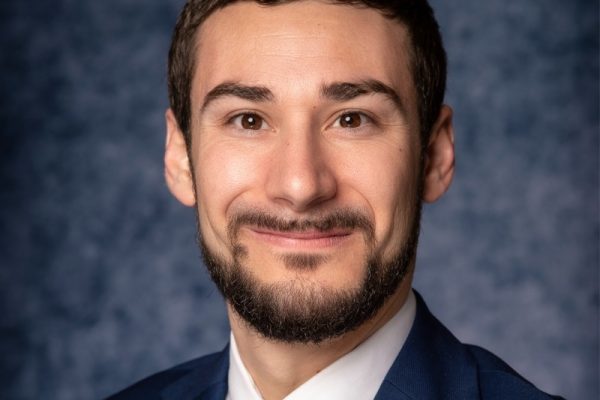 A man with a beard and short hair smiles at the camera, wearing a dark suit and a light-colored shirt with a tie. The background is a textured blue