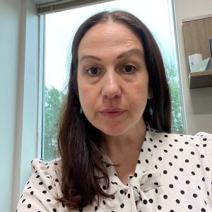 A woman with long, dark hair is sitting in an office setting. She is wearing a white blouse with black polka dots and has earrings. The background shows a window with a view of trees outside