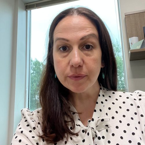 A woman with long, dark hair is sitting in an office setting. She is wearing a white blouse with black polka dots and has earrings. The background shows a window with a view of trees outside