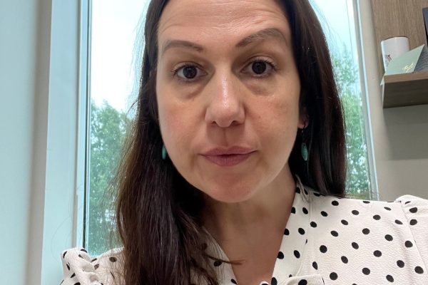 A woman with long, dark hair is sitting in an office setting. She is wearing a white blouse with black polka dots and has earrings. The background shows a window with a view of trees outside