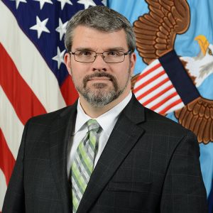 A man with short gray hair and glasses stands in front of a backdrop featuring the American flag and an eagle emblem. He is wearing a dark suit with a light green and white striped tie