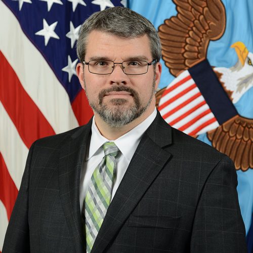 A man with short gray hair and glasses stands in front of a backdrop featuring the American flag and an eagle emblem. He is wearing a dark suit with a light green and white striped tie