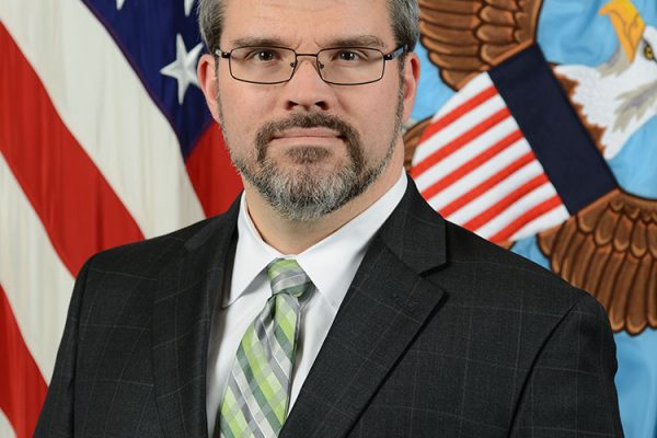 A man with short gray hair and glasses stands in front of a backdrop featuring the American flag and an eagle emblem. He is wearing a dark suit with a light green and white striped tie
