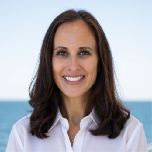 A woman with long, dark brown hair smiles at the camera, wearing a white button-up shirt. The background features a calm ocean and clear sky