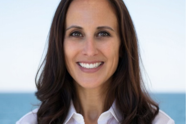 A woman with long, dark brown hair smiles at the camera, wearing a white button-up shirt. The background features a calm ocean and clear sky