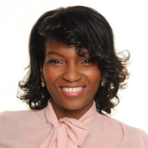 A woman with shoulder-length, curly black hair smiles warmly. She is wearing a light pink blouse with a bow tie at the collar and has earrings. The background is plain and light-colored