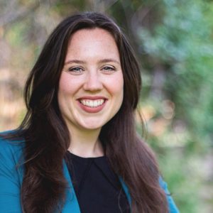A woman with long brown hair smiles at the camera, wearing a teal blazer over a black top. The background features blurred greenery, suggesting an outdoor setting