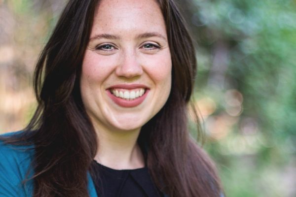 A woman with long brown hair smiles at the camera, wearing a teal blazer over a black top. The background features blurred greenery, suggesting an outdoor setting