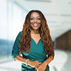 A woman with long, curly dreadlocks smiles while wearing a green blouse. She stands in a well-lit, modern interior space with large windows in the background