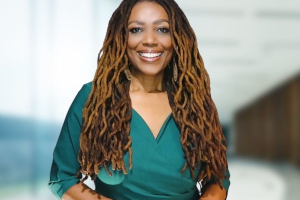 A woman with long, curly dreadlocks smiles while wearing a green blouse. She stands in a well-lit, modern interior space with large windows in the background