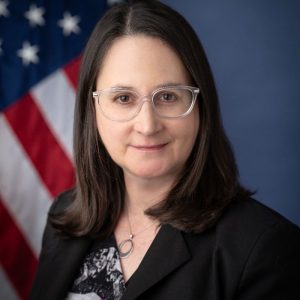 A woman with long brown hair and glasses is smiling at the camera. She is wearing a black blazer over a patterned top. In the background, there is an American flag
