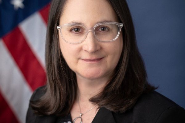 A woman with long brown hair and glasses is smiling at the camera. She is wearing a black blazer over a patterned top. In the background, there is an American flag