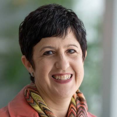 A woman with short, dark hair smiles warmly. She is wearing a colorful, patterned scarf and a light-colored jacket. The background is softly blurred, suggesting an indoor setting with natural light