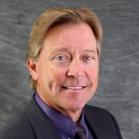 A man with light brown hair and a friendly smile is wearing a dark suit and a purple shirt. He is facing slightly to the right against a textured gray background
