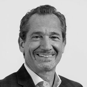A man with short, wavy hair and a slight smile is wearing a dark blazer over a light-colored shirt. He has a well-groomed beard and is facing the camera against a plain background