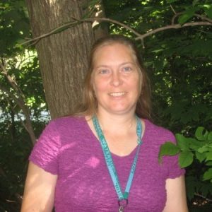 A woman with long brown hair, wearing a purple shirt and a teal lanyard, smiles while standing in front of a tree surrounded by green foliage