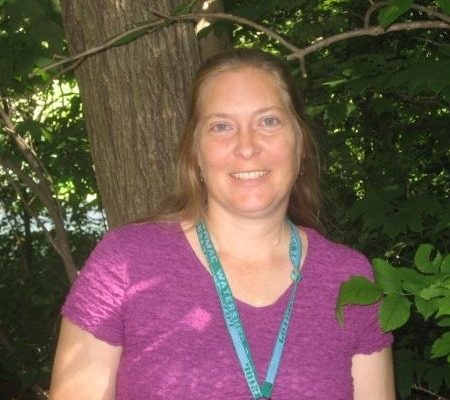 A woman with long brown hair, wearing a purple shirt and a teal lanyard, smiles while standing in front of a tree surrounded by green foliage