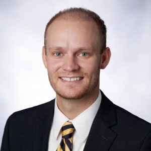 A man with short light brown hair and a beard smiles at the camera. He is wearing a dark suit with a white shirt and a striped tie. The background is a soft, light color