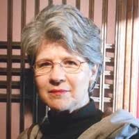 A woman with short gray hair and glasses is looking directly at the camera. She is wearing a black turtleneck and has a neutral expression. The background features wooden paneling