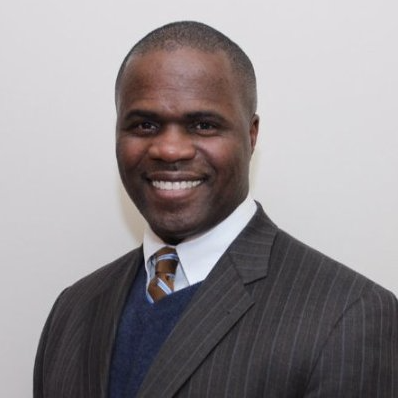 A man with short, closely cropped hair smiles while wearing a dark suit with a striped pattern, a white dress shirt, and a brown and white striped tie. He stands against a plain light-colored background