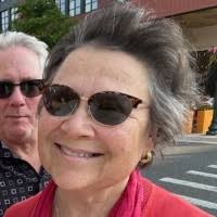 A woman with short, gray hair and sunglasses smiles at the camera. She is wearing a red top and has a scarf around her neck. In the background, a man with gray hair is partially visible