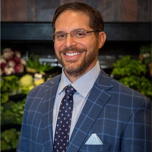A man with glasses and a beard smiles while wearing a blue plaid suit jacket, a light blue dress shirt, and a patterned tie. He stands in front of a display of fresh produce, including leafy greens and vegetables