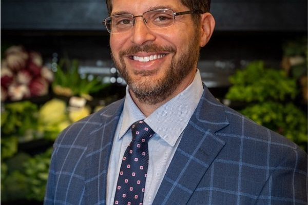 A man with glasses and a beard smiles while wearing a blue plaid suit jacket, a light blue dress shirt, and a patterned tie. He stands in front of a display of fresh produce, including leafy greens and vegetables