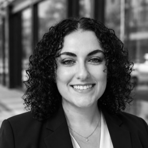 A woman with curly black hair smiles at the camera, wearing a black blazer over a white top. She stands in front of a building with large windows