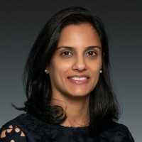 A woman with long, dark hair and a slight smile is wearing a navy blue top with a lace pattern. She has pearl earrings and is set against a dark gray background