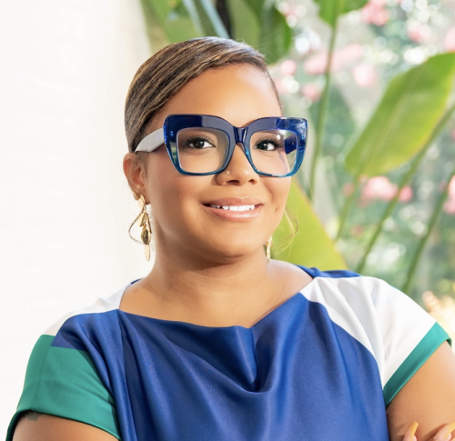 A woman with short, styled hair wears large, blue-framed glasses and a colorful top. She is smiling and posed in front of green plants with soft pink flowers in the background