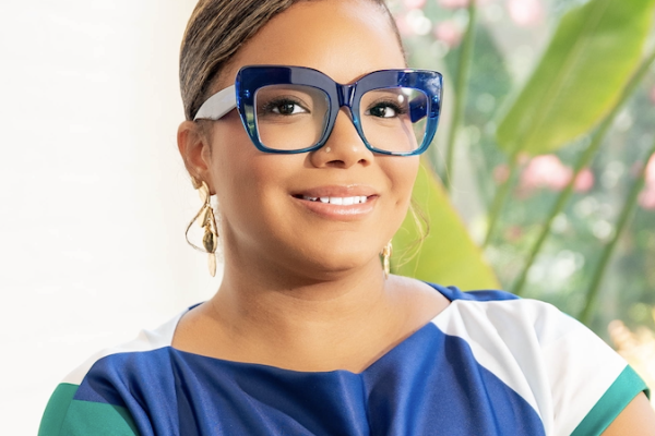 A woman with short, styled hair wears large, blue-framed glasses and a colorful top. She is smiling and posed in front of green plants with soft pink flowers in the background