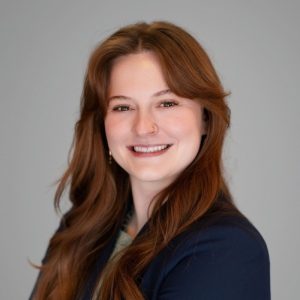 A young woman with long, wavy brown hair smiles confidently. She is wearing a dark blazer over a light-colored top and has a nose ring. The background is a neutral gray