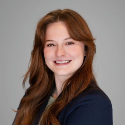 A young woman with long, wavy brown hair smiles confidently. She is wearing a dark blazer over a light-colored top and has a nose ring. The background is a neutral gray