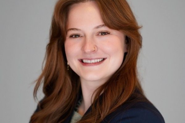 A young woman with long, wavy brown hair smiles confidently. She is wearing a dark blazer over a light-colored top and has a nose ring. The background is a neutral gray