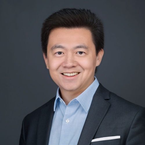 A man with short black hair smiles while wearing a dark suit jacket over a light blue shirt. The background is a solid dark color