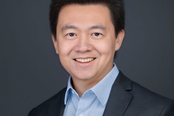 A man with short black hair smiles while wearing a dark suit jacket over a light blue shirt. The background is a solid dark color