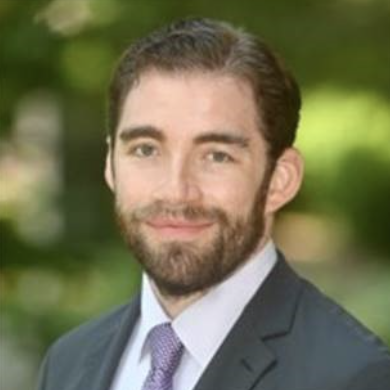 A man with a beard and short hair smiles while wearing a dark suit and a light purple tie. The background features greenery, suggesting an outdoor setting