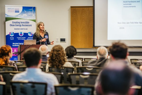 A woman stands at a podium, speaking to an audience during a presentation. She gestures towards a screen displaying a calendar and text related to clean energy businesses. In the background, a banner promoting Maryland Energy Innovation is visible. The audience, seated and facing her, appears engaged in the discussion