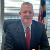 A man in a black suit and red tie sits at a desk, smiling. He has short, gray hair and is positioned in front of a window with blinds. A Maryland state flag is visible in the background