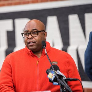 A man wearing glasses and an orange jacket speaks at a podium with microphones in front of a large banner. He appears to be addressing an audience during an event