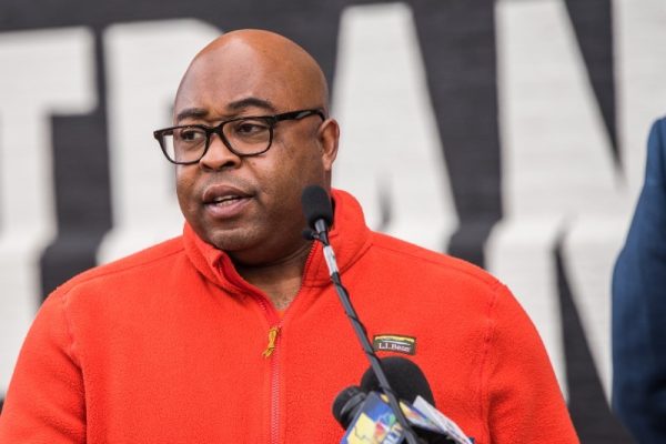 A man wearing glasses and an orange jacket speaks at a podium with microphones in front of a large banner. He appears to be addressing an audience during an event
