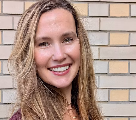A woman with long, wavy brown hair smiles at the camera. She is wearing a maroon top and has a gold necklace. The background features a light-colored brick wall