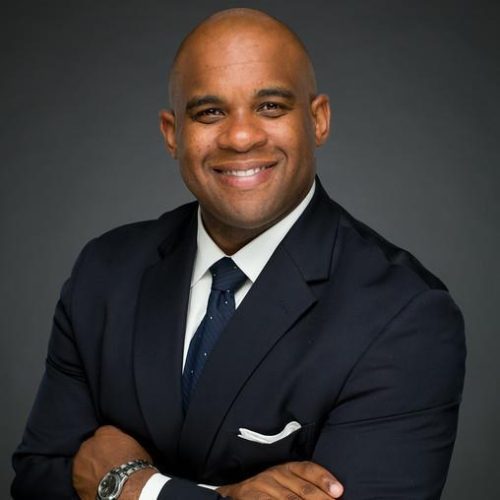 A man with a shaved head smiles confidently while wearing a dark suit and tie. He has his arms crossed and is positioned against a dark background