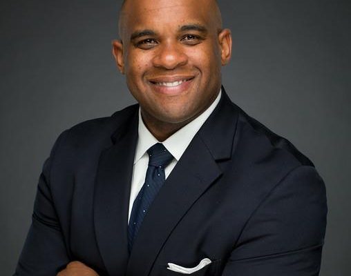 A man with a shaved head smiles confidently while wearing a dark suit and tie. He has his arms crossed and is positioned against a dark background