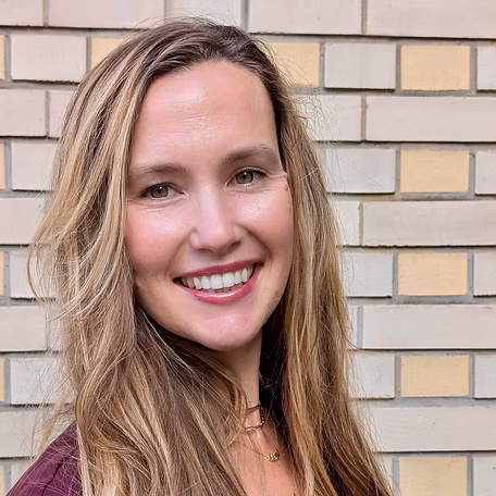 A woman with long, wavy brown hair smiles at the camera. She is wearing a maroon top and has a gold necklace. The background features a light-colored brick wall