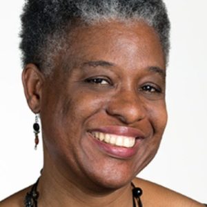 A smiling woman with short gray hair and earrings, wearing a black necklace. She has a warm expression and is facing the camera