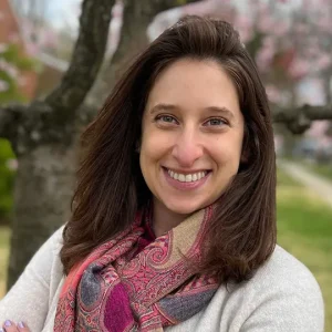 A woman with long brown hair smiles at the camera, wearing a light sweater and a colorful scarf. She stands outdoors with blurred trees and pink blossoms in the background