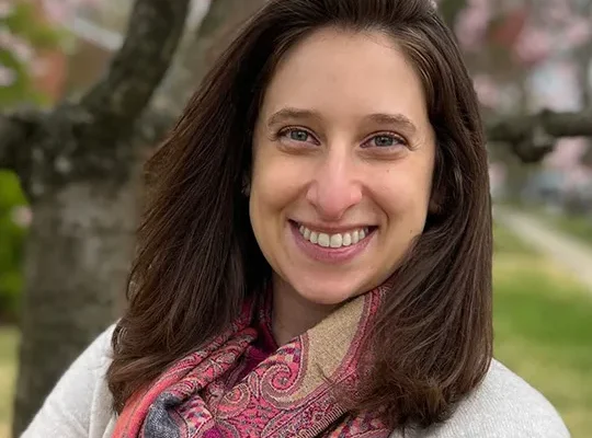 A woman with long brown hair smiles at the camera, wearing a light sweater and a colorful scarf. She stands outdoors with blurred trees and pink blossoms in the background
